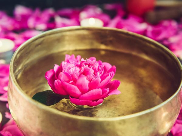 Tibetan singing bowl with floating in water pink peony flower. Burning candles and petals on the black stone background. Meditation and Relax. Exotic massage. Vertical. Selective focus. Copy space. Tibetan singing bowl with floating in water pink peony flower. Burning candles and petals on the black stone background. Meditation and Relax. Exotic massage. Vertical. Selective focus. Copy space
